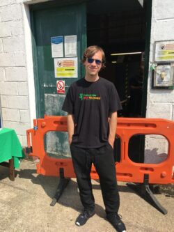 Oleg is standing up and facing forward to the camera wearing his black Tools for Self Reliance t-shirt, black trousers and sunglasses in front of the workshop back door. There is a also a plastic orange barrier behind him.