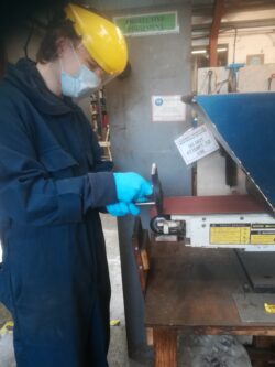 Oleg is standing sign on in a navy blue boiler suit with mas, visor and blue gloves at a tool sharpening machine at the Netley Marsh workshop