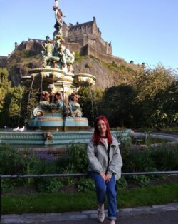 Alfiia is sat on the railing infront of the The Ross Fountain, an ornate fountain in the city of Edinburgh. In the background is Edinburgh Castle. She is wearing a light grey jacket and blue jeans.