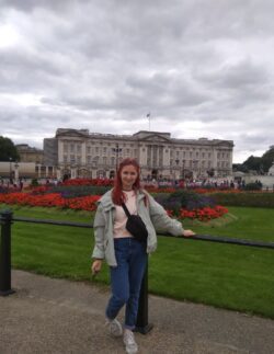 Alfiia is stood in the middle of the foreground wearing blue jeans, a pale pink jumper, a light grey jacket and a bag worn across her front. Leaning on black railings in front of a lawn with a red flower arrangement behind and Buckingham Palace in the background. 
